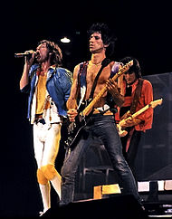 The Rolling Stones på scen i december 1981. Från vänster: Mick Jagger i blå jacka med gula kläder och ett svart bälte som sjunger i en mikrofon, Keith Richards klädd i svarta byxor och en liten lila väst (ingen skjorta) spelar en svart gitarr till vänster – och lite framför – Jagger, Ronnie Wood klädd i orange jacka och svart skjorta/byxor spelar en beige gitarr bakom Jagger och Richards.