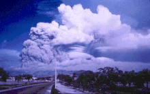 Zdjęcie przedstawia krajobraz. Na pierwszym planie pusta droga i tropikalna roślinność. W oddali zajmujące większość zdjęcia kłęby dymu i chmur, z widocznymi kilkoma mniejszymi chmurami w kształcie soczewek.