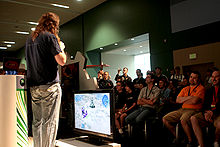 Um homem vestindo uma camisa preta e jeans e com um microfone na mão fala para uma multidão sentada. No centro do quadro está uma televisão de tela plana exibindo a jogabilidade de Halo Wars.