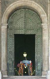 Frontale Farbfotografie von einem grossen grünen Toreingang mit zwei Gardisten. Der rechte Gardist steht seitlich mit einer Hellebarde. Im Innern des Gebäudes sind Deckenlampen zu sehen.