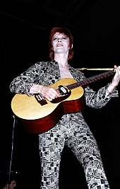 Una fotografia a colori di David Bowie con una chitarra acustica