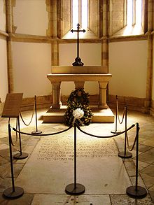 Foto dari kapel kecil yang memuat sebuah lempengan batu berbatas-tali terpasang dalam lantai serta sebuah altar batu pada bagian latar dengan karangan bunga dan salib logam besar