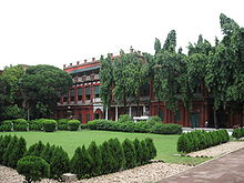 A brick-red mansion in the background, shaded by a row of large trees; in the foreground, a manicured lawn with a perimeter of trimmed round bushes.