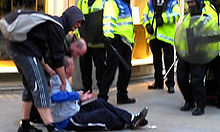 Tomlinson sitting on the ground facing the police with his arms outstretched; two men are helping him up