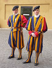 Frontale Farbfotografie von zwei Männern in Gardeuniform, die vor einer ockerfarbenen Wand stehen. Die Uniform hat blau-gelb längsgestreifte Puffärmel und Pumphosen mit rotem Stoff in den Falten. Der untere Beinstoff liegt eng am Bein und ist ebenfalls blau-gelb gestreift. Der Stehkragen ist weiss und sie tragen eine schwarze Baskenmütze.