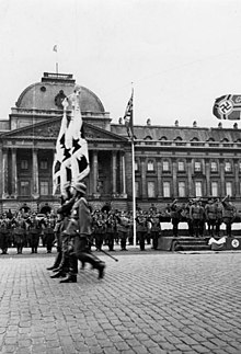 Photographie en noir et blanc montrant des soldats alignés au premier plan, vus de profil, et le palais royal à l'arrière-plan, un drapeau nazi flottant dans les airs en haut à droite du cadre.
