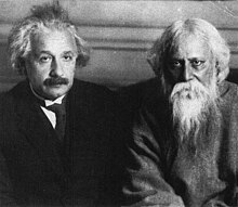 A moustached man in a lounge suit and necktie (left) sits next to a white-haired, bearded man dressed in robes (right). Both look toward the camera.