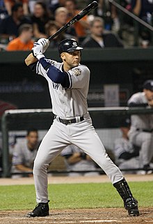 A man in a gray baseball jersey and a navy blue batting helmet stands in a batters box, preparing to swing at a pitch.