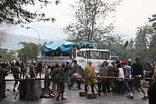 Ein mit UN beschrifteter Lastwagen auf dessen Ladefläche sich unter einer Plane eng gedrängte Menschen befinden, steht auf einer asphaltierten Straße. Er ist von einigen bewaffneten Blauhelmsoldaten umgeben sowie Gruppen von Ruandern, die teils Macheten in Richtung des Lastwagens erheben. Vor und hinter dem Lastwagen sind Absperrungen. Rechts im Bild stehen Mitarbeiter der Filmcrew.