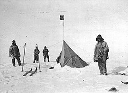 Fire skikkelser i tunge klær står nær et spisst telt med et lite, firkantet flagg. Den omkringliggende bakken er islagt. Ski og staver er vist til venstre.