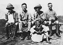 Four men in khaki military uniforms, two wearing pith helmets (one of whom has his arm in a sling), seated in front of a boy