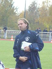 Jean-Guy Wallemme en tenue d'entraîneur, avec des carnets dans la main.