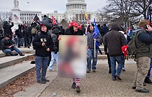 A Trump supporter carries a QAnon-tagged placard with Jesus wearing a MAGA hat at the moment the U.S. Congress was violently attacked by rioters on January 6, 2021. (The placard is blurred for copyright reasons.)