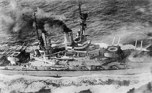 Overhead view of a large battleship; black smoke pours from its smoke stacks as it steams through choppy seas