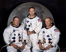 Tiga astronaut dalam setelan antariksa tanpa helm duduk di depan foto Bulan.