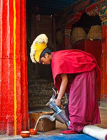 Monk in red robes pouring butter tea into a bowl in the ground from a large silver-colored tea pot