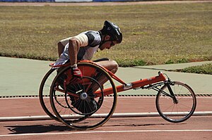 Um atleta, usando óculos escuros e capacete, movimenta uma cadeira de rodas numa pista de atletismo