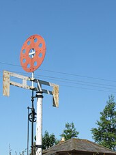 Un poste blanco soporta dos brazos cruzados unidos por cuatro barras verticales cortas para formar una cuadrícula. Arriba hay un disco rojo perforado por ocho agujeros.