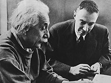 Einstein menulis di meja. Oppenheimer duduk di sebelahnya.