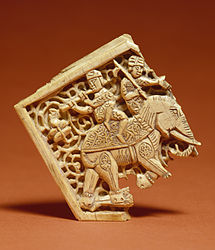 Dues figures, una probablement músic, condueixen un elefant; part dels accessoris amb incrustacions d'una porta o un moble gran.