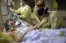 Health care providers attending to a person on a stretcher with a gunshot wound to the face. The patient is intubated, and a mechanical ventilator is visible in the background.