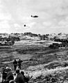 Okinawa's Landscape in the south is marked by fields of grain and vegetables, broken only by humps of coral, farmhouses, and villages. Navy plane flying over such terrain is shown dropping supplies to the last fast-moving American troops early in the campaign