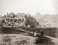 Sepia photograph of a ruined mountain fortress behind fields and farmers.