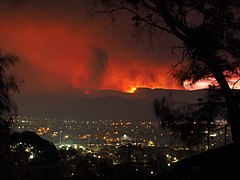 Fotografie večera v údolnej osade. Obzor v kopcoch za ňou je červeno osvetlený od požiarov.