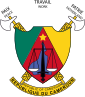Tricolor shield before two crossed fasces. Its center is an inverted red kite shape covered with a purple outline of Cameroon below a gold star, with the scales of justice superimposed. Its left is green and its right is gold. Banners with fine print are above and below.