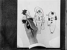 Photographie en noir et blanc d'une main, vue de dessus, dessinant trois personnages sur une feuille de papier.