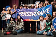 Cerca de vinte alunos finalistas do ensino fundamental estão de pé em um palco, todos sorrindo, e segurando uma faixa que diz "Rover de perseverança da NASA". Na frente deles, no palco, está um veículo espacial em miniatura.