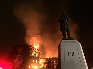 This photo shows a large, tall building, the National Museum of Brazil, engulfed in flames. To the left of the image, the red glow of fire-truck lights can be seen. In the foreground, to the right, there is a statue of Emperor Pedro II, the name "Pedro II" engraved on its base.