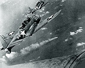 černobílá fotografie dvou nad mořem letících jednomotorových bombardovacích letounů. Několik set metrů pod nimi je hořící loď, za kterou se táhne vlečka kouře.