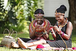 Ruohikolla käsitöitä langoista valmistavat kaksi kenialaista naista, joilla on päällään perinteisiä koruja.