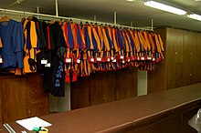 Farbfotografie von vielen Uniformen mit Namensschildern, die an einer Stange hängen. Im Hintergrund und rechts sind Schränke zu sehen. Im Vordergrund steht ein langer Ausgabetresen mit Papier und Markern.