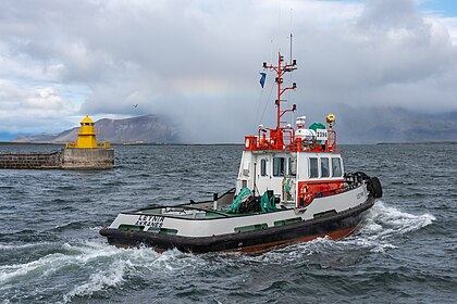 Remorkér Leynir mířící z Reykjavíku do zálivu Faxaflói, s horským hřbetem Esjan v pozadí