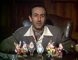 A color photograph of Walt Disney sitting behind a desk. Seven figurines are standing before him.