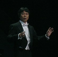 Photo d'un Japonais en costume de cérémonie noir sur scène de fond noir également.