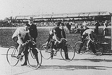Photo de trois cyclistes sur une piste devant la ligne de départ, avec trois hommes leur aidant à rester en équilibre.
