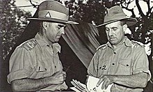 Two men in military uniform conferring in front of a tent. The one on the right is holding a stack of papers.