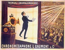 Illustration of a theater from the rear right of the stage. At the front of the stage a screen hangs down with the projected image of a tuxedoed man holding up a text and performing. In the foreground is a gramophone with two horns. In the background, a large audience is seated at orchestra level and on several balconies. The words "Chronomégaphone" and "Gaumont" appear at both the bottom of the illustration and, in reverse, at the top of the projection screen.