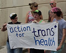Trois personnes tenant une banderole sur laquelle est écrite Action for trans health.