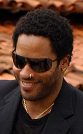 Lenny Kravitz sorrindo com um smoking e óculos escuros.