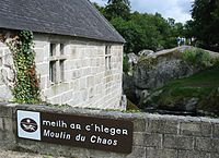 Side of a stone building next to a stream; low stone wall in the foreground has a sign reading Mill of Chaos in both Breton and French; Meilh ar C'hlegr and Moulin du Chaos