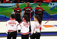 Quatro jogadoras da Rússia aparecem perfiladas em primeiro plano, de costas, usando uniformes brancos. Em segundo plano, quatro jogadoras do Canadá, também perfiladas e de costas, aparecem usando uniformes pretos