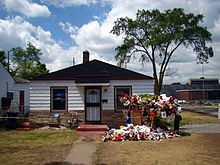The single-storey house has white walls, two windows, a central white door with a black door frame, and a black roof. In front of the house there is a walk way and multiple colored flowers and memorabilia.