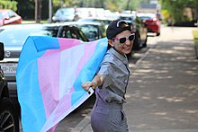 Jeune garçon trans dans la rue, tendant les bras comme Superman avec, en guise de cape, le drapeau trans
