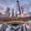 Illuminated water falls into 9/11 Memorial south pool at sunset, and glass-clad One World Trade Center with other skyscrapers in the background