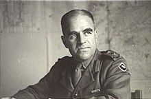 A man in military uniform sitting down and gazing off to the side. Maps are posted on the walls behind him.