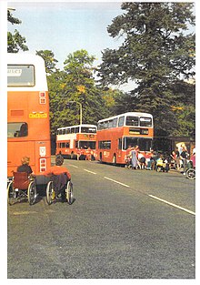 This colour photograph shows a road with three double decker buses stopped, each surrounded by protesting disabled people, some in wheelchairs. The buses are painted bright orange.
